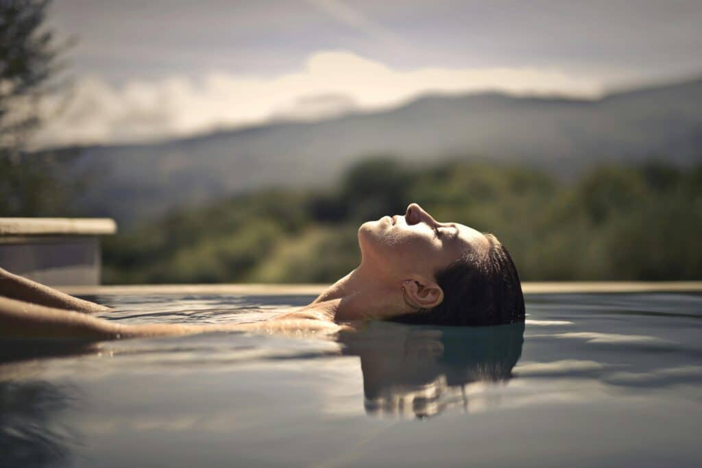 Femme flottant, les yeux fermés, dans l'eau d'une piscine à débordement ou d'un spa extérieur avec un paysage de montagne flou en arrière-plan, symbolisant la détente et le bien-être.