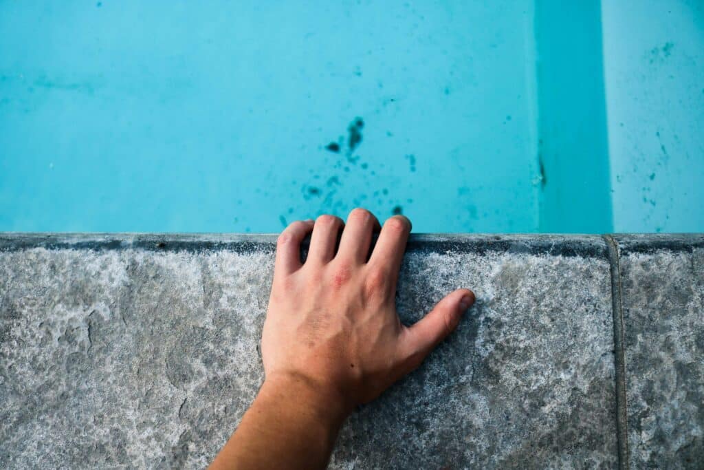 Main agrippée à la margelle en pierre d'une piscine dont l'eau est d'un bleu clair légèrement taché, illustrant la proximité avec la ligne d'eau et l'entretien des bords.